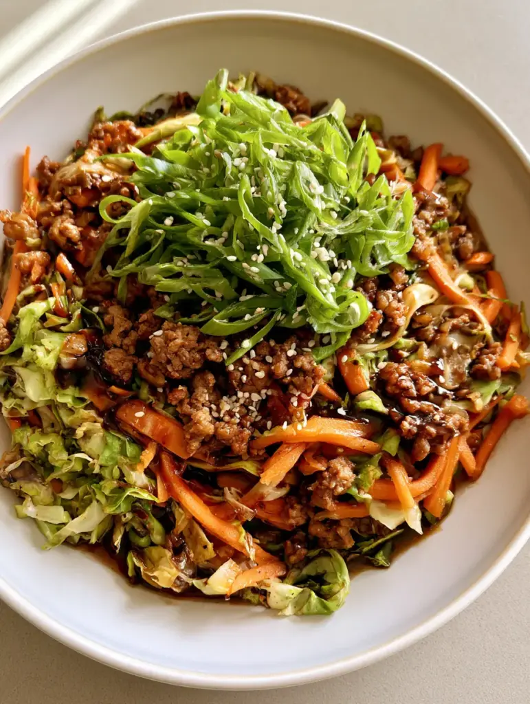 Egg Roll in a Bowl: The Ultimate 15-Minute One-Pan Dinner 1 A white bowl containing Egg Roll in a Bowl topped with white sesame seeds and bright green scallions on a light tabletop.